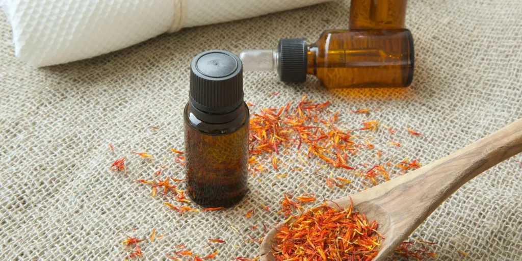 Small amber dropper bottle of natural oil with safflower petals and a wooden spoon on burlap.